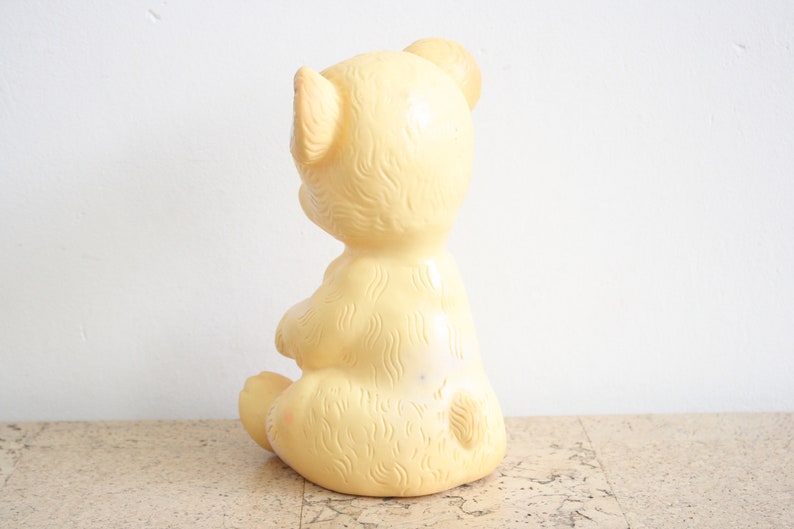 May include: A yellow rubber teddy bear toy sitting on a cork surface. The bear has a simple design with a smooth, rounded body and a small, pointed nose.