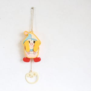 May include: A vintage, hanging baby mobile featuring a cartoon clown design. The clown has a yellow body, red shoes, and a blue and yellow hat. A white string and a decorative, off-white, heart-shaped pull are attached.