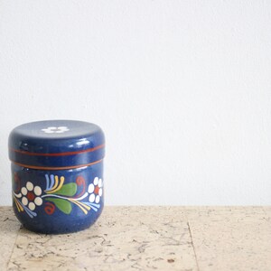 May include: A blue ceramic jar with a lid, decorated with white flowers and green leaves. The jar has a red stripe around the top.