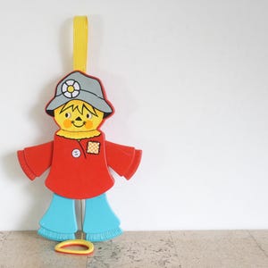 May include: A colorful, vintage scarecrow decoration. The scarecrow has a yellow face, a gray hat, and a red shirt and arms. It has blue pants and a yellow loop at the bottom. A yellow ribbon is attached to the top.