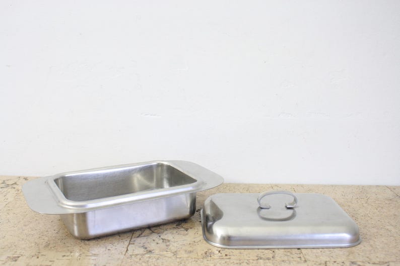 Puede incluir: Una mantequera rectangular de acero inoxidable con tapa a juego. El plato tiene un asa a cada lado y la tapa tiene un asa curva. La mantequera est&aacute; abierta y la tapa est&aacute; colocada al lado.