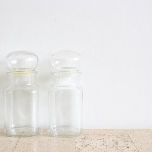 May include: Two clear glass jars with round lids. The jars are empty and have a square shape.