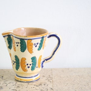Puede incluir: Una jarra de cerámica blanca con asa azul y un patrón floral colorido en tonos de verde, amarillo y azul.