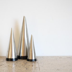 May include: Three silver-colored, cone-shaped decorative objects with black bases. The objects are arranged in a group, varying in size, against a white background. The objects are made of metal and have a sleek, modern design.