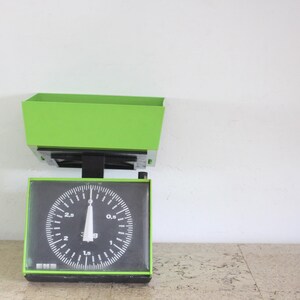 May include: A vintage mechanical kitchen scale featuring a bright lime green metal housing and a large rectangular tray. The front display has a black face with white circular markings, numbers ranging from 0 to 3 kg, and a single white needle.