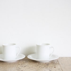 以下が含まれることがあります： リブ模様の白いセラミック製のコーヒーカップ2つと、それに合うソーサー。カップとソーサーは薄い茶色の表面の上に置かれています。