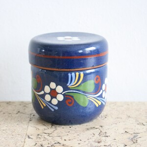 May include: A blue ceramic jar with a lid, decorated with white flowers and green leaves. The jar has a red rim and a white flower on the lid.
