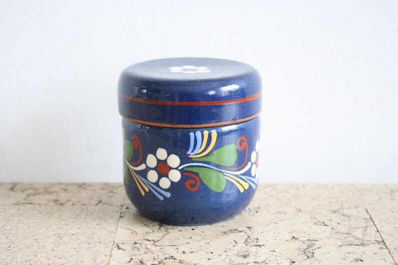 May include: A blue ceramic jar with a lid, decorated with white flowers and green leaves. The jar has a red rim and a white flower on the lid.