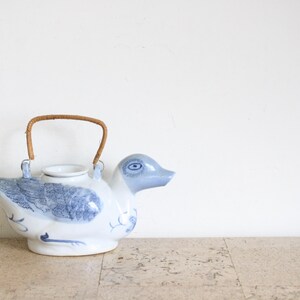 May include: A white and blue ceramic teapot shaped like a duck. The teapot has a woven handle and blue painted details on the body and head. The teapot is sitting on a light-coloured surface.