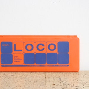 Puede incluir: Una caja de plástico naranja con la palabra "mini LOCO" en letras azules. La caja también tiene cuadrados azules y texto en holandés. La caja está cerrada y colocada contra una pared blanca.