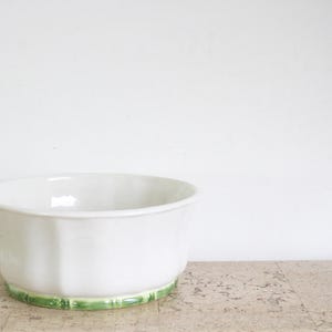 May include: A white ceramic bowl with a fluted design and a green base. The bowl has a wide, open top and sits on a light-coloured surface against a white background. This decorative bowl is suitable for various uses.