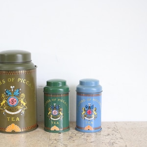 Può includere: Tre barattoli di tè vintage in verde oliva, verde foresta e azzurro. Ogni barattolo presenta un stemma decorativo e la scritta "SONS OF PICCADILLY TEA". I barattoli sono cilindrici con coperchi abbinati.