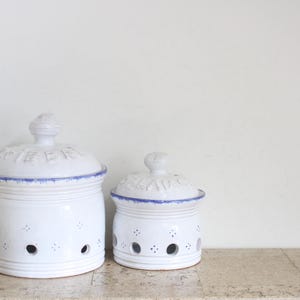 Op de afbeelding: Twee witte keramische voorraadpotten met deksels, voorzien van blauwe randen en reliëfletters op de deksels. De cilindrische lichamen hebben een reeks ronde ventilatiegaten aan de basis en kleine decoratieve stippenpatronen.