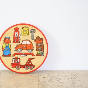 Könnte beinhalten: Ein Vintage-Holzpuzzle mit kreisförmigem Design und rotem Rand. Das Puzzle zeigt ein Autoreparatur-Thema mit Cartoonfiguren, einem Auto, einem Abschleppwagen und anderen verwandten Objekten. Die Farben sind hauptsächlich Rot, Orange, Gelb und Blau.
