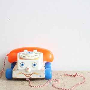 Può includere: Un telefono giocattolo vintage con cornetta arancione e ruote blu. Il corpo bianco presenta un volto sorridente e un quadrante rotante. Il telefono è collegato a un cavo a strisce rosse e bianche, evocando nostalgia.