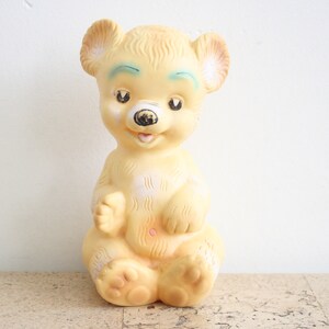 May include: A vintage yellow rubber toy bear with painted blue eyes and a black nose. The bear is sitting with its paws in front of it.