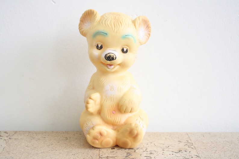 May include: A vintage yellow rubber toy bear with painted blue eyes and a black nose. The bear is sitting with its paws in front of it.