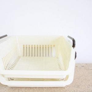Puede incluir: Una cesta de almacenamiento de plástico blanco con asas marrones. La cesta tiene un patrón de rejilla y está diseñada para apilarse.