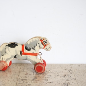 Può includere: Un giocattolo da trainare in legno vintage a forma di cavallo con corpo color crema, criniera e coda grigie e macchie nere. Il cavallo presenta una pettorina rosso brillante e quattro ruote in legno rosse fissate alle zampe con metallo.