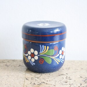May include: A blue ceramic jar with a lid, decorated with white flowers and green leaves. The jar has a brown rim and a white flower on the lid.
