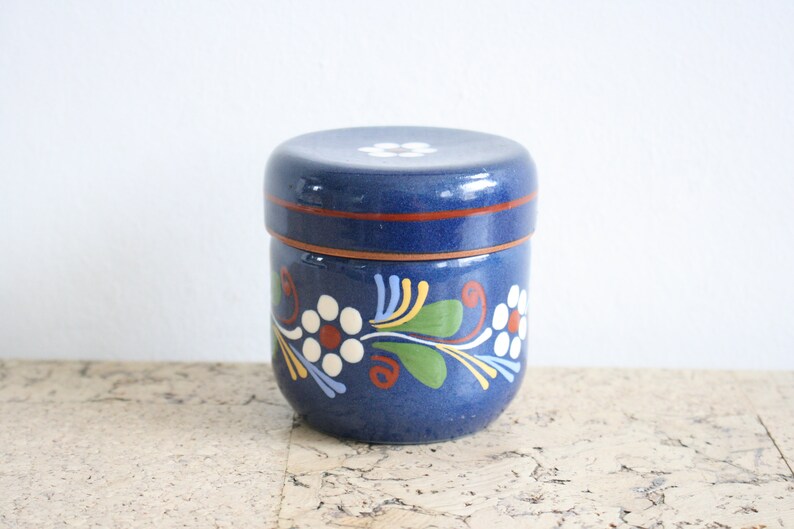 May include: A blue ceramic jar with a lid, decorated with white flowers and green leaves. The jar has a brown rim and a white flower on the lid.