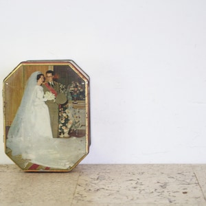 Puede incluir: Una caja de hojalata vintage con un diseño octogonal en relieve. La caja presenta una ilustración colorida de una novia y un novio con su atuendo de boda. La novia lleva un vestido blanco con un velo largo, y el novio lleva un traje oscuro. La pareja está de pie frente a un arreglo floral.