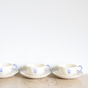 May include: Three white teacups with blue floral designs and matching saucers. The cups and saucers are arranged in a row on a light beige surface.