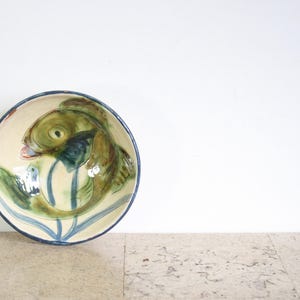 Peut inclure: Un bol rond en céramique avec un motif de poisson peint à la main. Le poisson est représenté dans des tons de vert, de bleu et de marron sur un fond crème. Le bord du bol est bleu foncé. Le bol est posé sur une surface texturée.