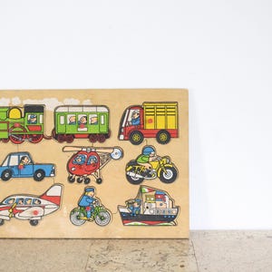 Op de afbeelding: Een houten puzzelbord met verschillende voertuigen, waaronder een trein, een vrachtwagen, een auto, een helikopter, een motorfiets, een vliegtuig en een boot. De voertuigen zijn in felle kleuren geverfd en hebben cartoonfiguren.