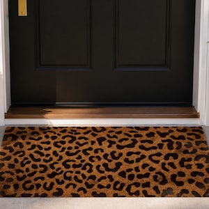 Può includere: Uno zerbino rettangolare con un motivo leopardato marrone e nero. Il tappetino ha una consistenza fibrosa e ruvida ed è posizionato orizzontalmente su un pavimento piastrellato chiaro davanti a una porta nera con cornice bianca.