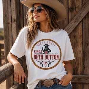 Könnte beinhalten: Ein weißes kurzärmeliges T-Shirt mit einem kreisförmigen Aufdruck, der von einem braunen Seil umrandet ist. Das Design zeigt die Silhouette einer Person mit Cowboyhut, eine rote Rose, zwei Revolver, einen Rinderschädel und den Text KINDA SWEET KINDA BETH DUTTON.