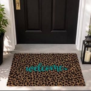 Può includere: Uno zerbino rettangolare con un motivo leopardato marrone e nero. La parola welcome è scritta al centro in un carattere corsivo color verde acqua, seguita da un piccolo simbolo a forma di cuore dello stesso colore alla fine del testo.