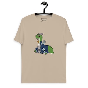 May include: A beige t-shirt with a cartoon dinosaur character wearing a blue and white outfit and a silver helmet. The dinosaur is green with a yellow beak. The shirt is hanging on a black hanger.