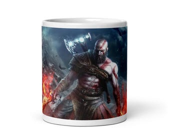 May include: White ceramic mug featuring a detailed illustration of a muscular warrior with a beard, axe, and red markings. The background shows a fiery scene with a dark, stormy sky. The mug is a standard size.