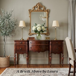 May include: A dark wood sideboard with two drawers and a central cabinet, topped with two lamps and a gold-framed mirror. A vase of white flowers sits on the sideboard. The text "A Brush Above by Laura" is at the bottom.