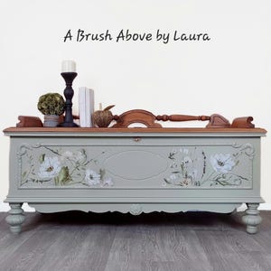May include: A Brush Above by Laura. A vintage-style chest painted in a soft green with floral accents. The chest has a wooden top and decorative legs. A candle holder, books, and decorative items sit on top.