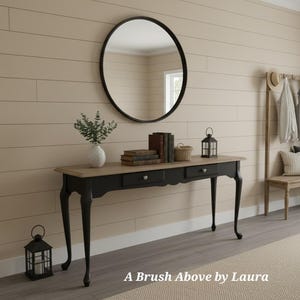 May include: A black and tan console table with three drawers, a round black mirror, and decorative items. The table has curved legs and a light-colored top. A black lantern and a coat rack are also visible.