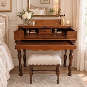 May include: A wooden vanity desk with a flip-top lid revealing interior storage compartments, a pull-out drawer, and four turned legs. The desk is paired with a rectangular, cream-colored upholstered stool featuring button tufting and nailhead trim.
