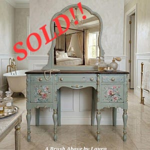 May include: A sage green vanity table with a matching arched mirror, featuring hand-painted floral motifs on the drawers and doors, gold-toned hardware, and slender turned legs. The text "SOLD!!" and "A Brush Above by Laura" are visible.