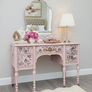 May include: A light pink vanity table featuring floral patterned drawer fronts, gold-toned hardware, and a central mirror. The text A Brush Above by Laura is visible at the top. The piece stands on four legs with casters and includes a matching mirror.