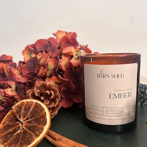 May include: A brown glass candle with a label reading "EMBER" from "The Barn Shed." The candle sits beside dried orange slices, cinnamon sticks, and burgundy flowers. The candle is handmade and poured in Yorkshire, England.