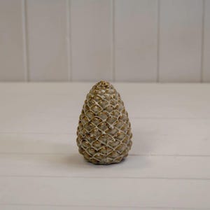May include: A ceramic pinecone ornament with a textured surface and gold detailing. The pinecone is a light brown colour and is displayed on a white surface. The background is a white wall with vertical wooden panels.