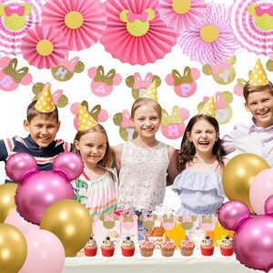 May include: Pink and gold Minnie Mouse themed birthday party decorations. The decorations include a banner, paper fans, hats, and balloons. The banner says "Happy Birthday".