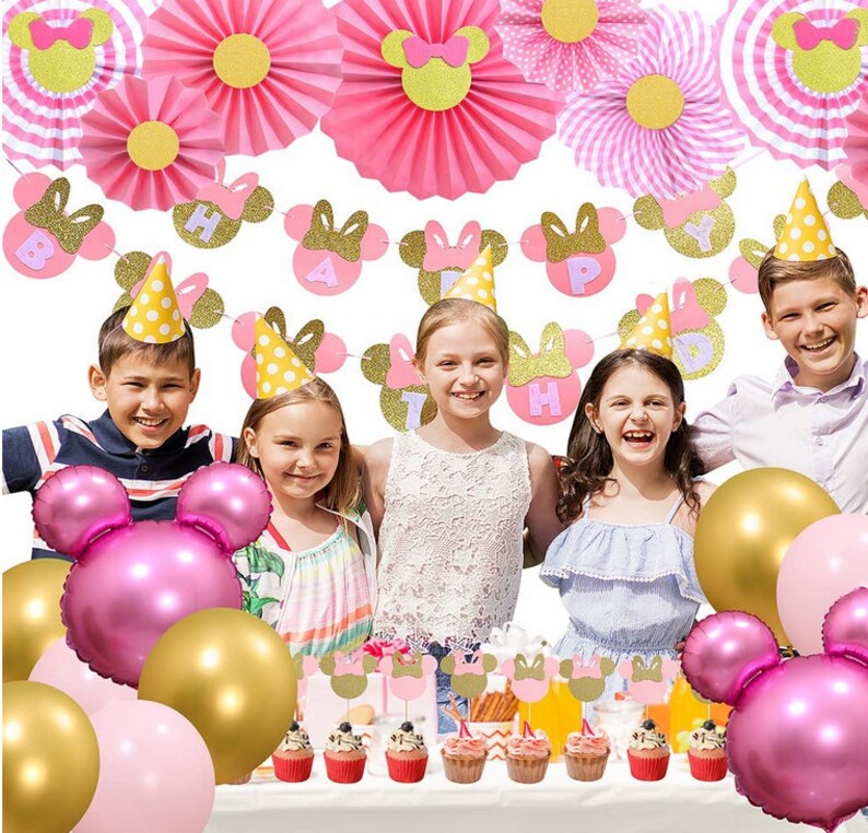 May include: Pink and gold Minnie Mouse themed birthday party decorations. The decorations include a banner, paper fans, hats, and balloons. The banner says "Happy Birthday".