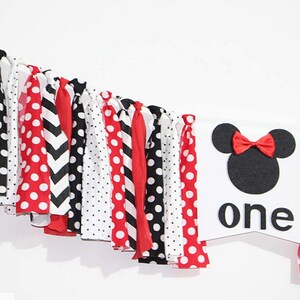 May include: A white banner with a black and red Minnie Mouse head and the word "one" in black letters. The banner is decorated with a red, black, and white fabric garland.