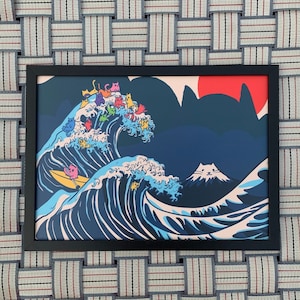 May include: Framed art print depicting cats surfing on ocean waves, with a red sun and a mountain in the background. The artwork is in a black frame and is displayed on a woven surface.