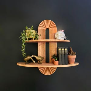 May include: A wooden shelf with a unique arch design, featuring two shelves. The top shelf has a small potted plant, a white skull, and a book. The bottom shelf has two golden bird figurines, a small potted plant, and three books. The shelf is mounted on a black wall.