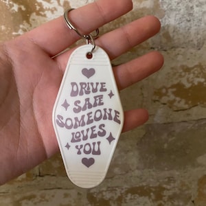 Könnte beinhalten: Weißer Motel-Schlüsselanhänger mit einem lila Herz und dem Text "Drive Safe Someone Loves You" in einer Retro-Schrift.