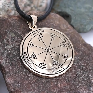 May include: Gold-colored pendant necklace with a black cord. The circular pendant features intricate engraved symbols and text around the edge and in the center. The pendant is displayed on a textured, dark stone surface.