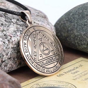 Puede incluir: Un colgante de color bronce con un diseño intrincado, con formas geométricas y texto. El colgante está en un collar de cordón negro. El fondo incluye una piedra y un papel con el texto "6th Pentacle of Sun".